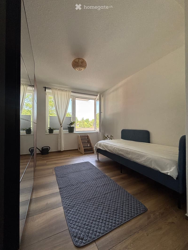 Bright bedroom with wooden flooring, a dark blue bed with white bedding, a grey quilted rug, a wicker hanging lamp, and a window with white curtains and potted plants on the sill.