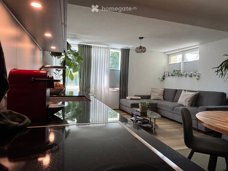 Modern living room with a grey sectional sofa, coffee table, wooden dining table, and pendant lighting. Large windows with curtains and plants add to the decor.