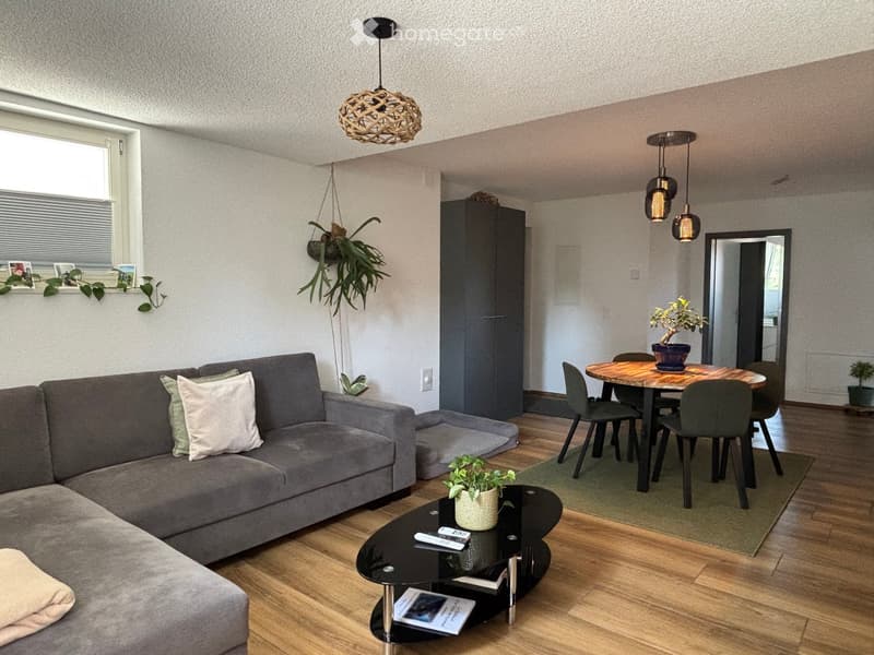 Modern living room with a grey sectional sofa, black coffee table, wooden dining table with chairs, pendant lights, and several potted plants.