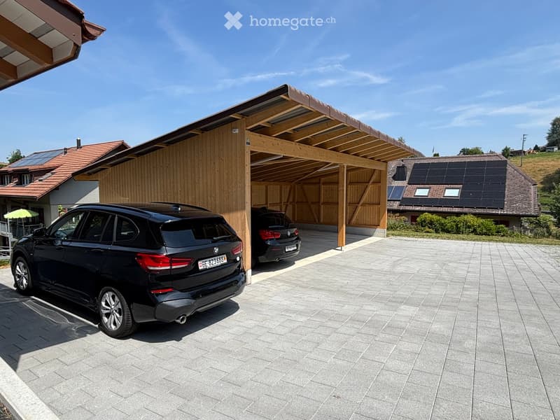 Outdoor area with a covered parking space for two cars, featuring a wooden carport and a paved driveway. Solar panels are installed on nearby buildings.