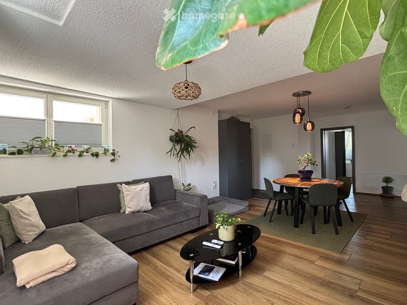 A modern living room with a grey sectional sofa, a black coffee table, a dining table with chairs, pendant lights, and several potted plants. The room features wooden flooring and large windows with blinds.