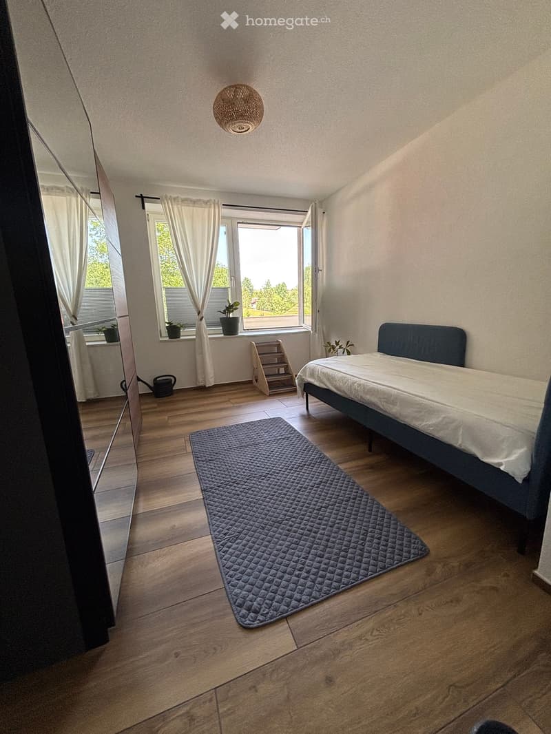 Cozy bedroom with wooden flooring, a double bed with white bedding, a blue quilted rug, a window with white curtains, and small potted plants on the windowsill.