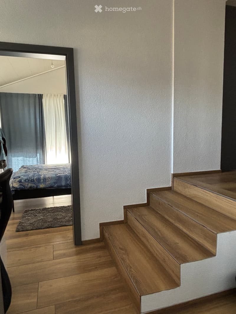 A modern hallway with wooden flooring, a large mirror, a staircase leading upwards, and a small bedroom visible in the reflection.