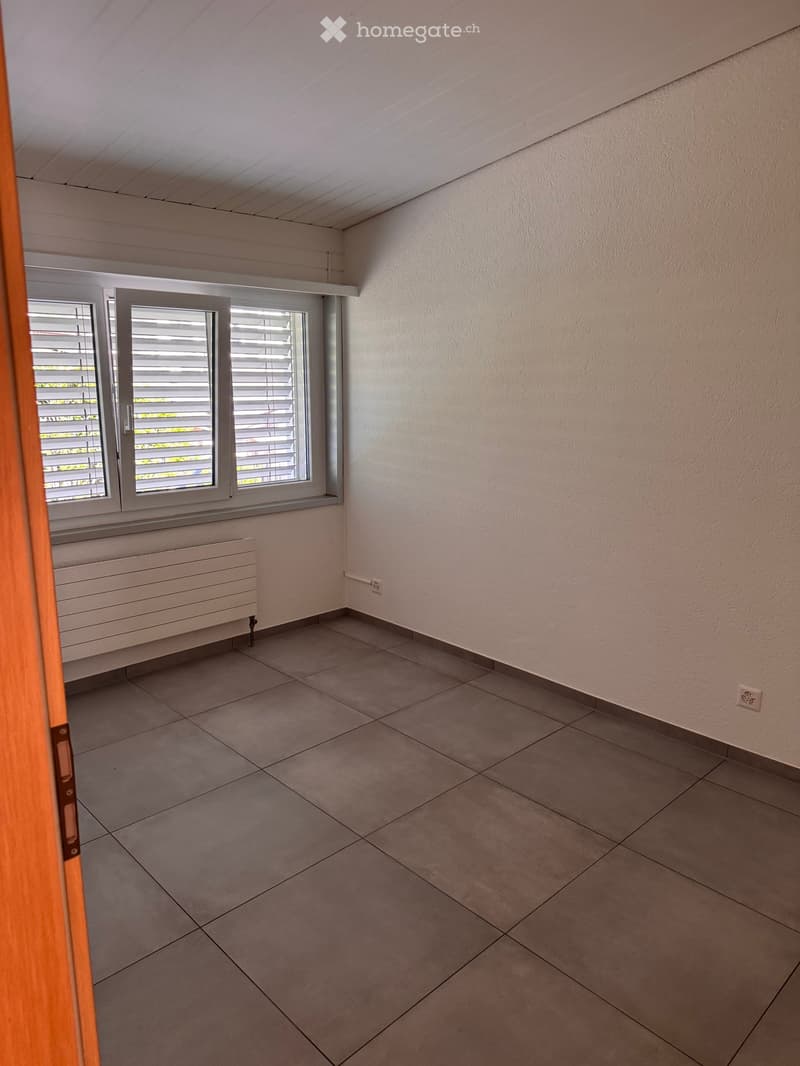 Empty room with tiled floor, white walls, and large windows with blinds. Radiator visible under windows.