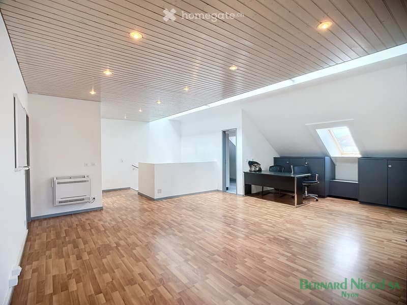Spacious attic or loft space with wooden flooring, white walls, and a slanted ceiling with recessed lighting. The room features a desk and office equipment, suggesting it could be used as a home office or study area.
