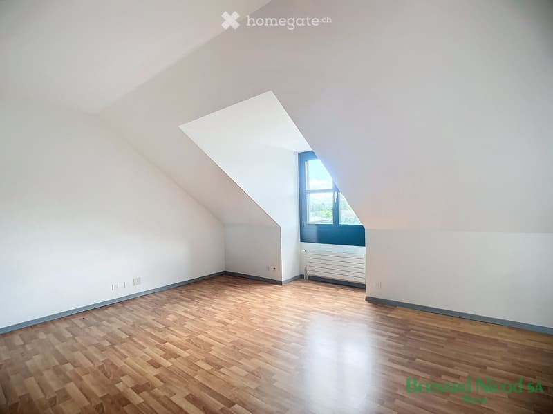 Spacious attic bedroom with hardwood flooring, white walls, and a large window providing natural light and a view.