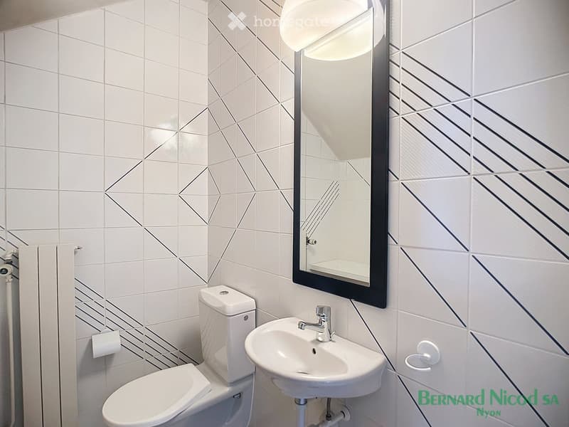Bathroom with white tiled walls featuring geometric patterns, a toilet, a sink with a mirror, and a light fixture.
