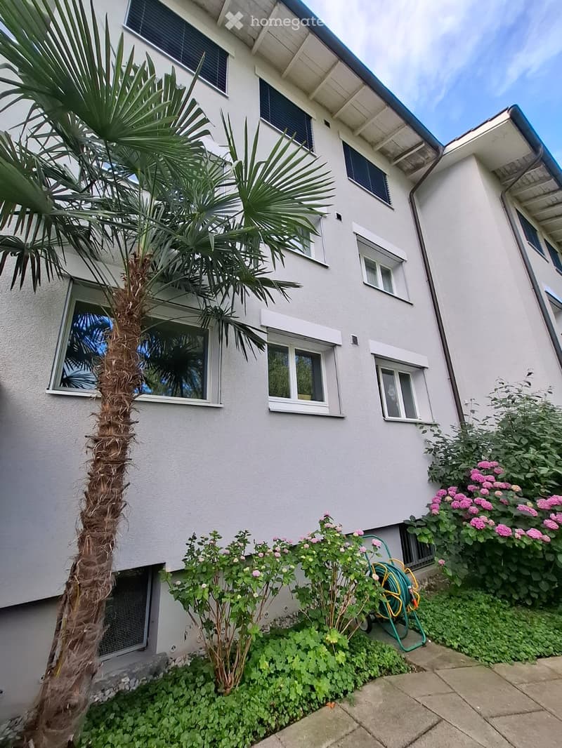 A two-story white building with multiple windows, surrounded by greenery including a palm tree and flowering bushes.