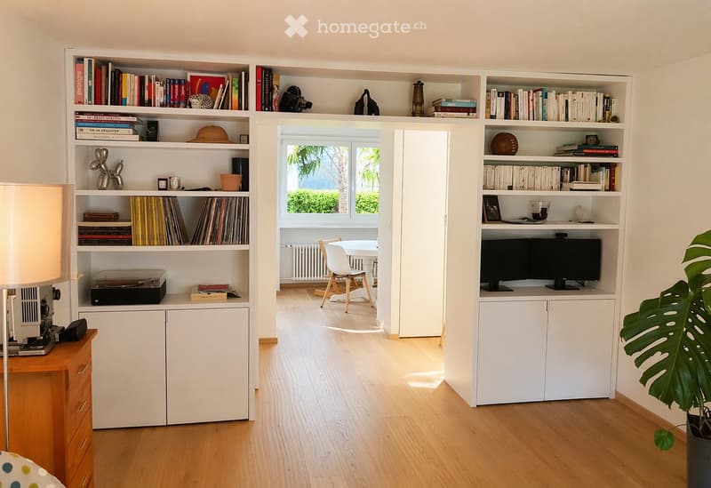 A modern, well-lit room with light wooden flooring, white built-in bookshelves filled with books and decorative items, a small table and chair in the adjacent space, and a potted plant near the corner.