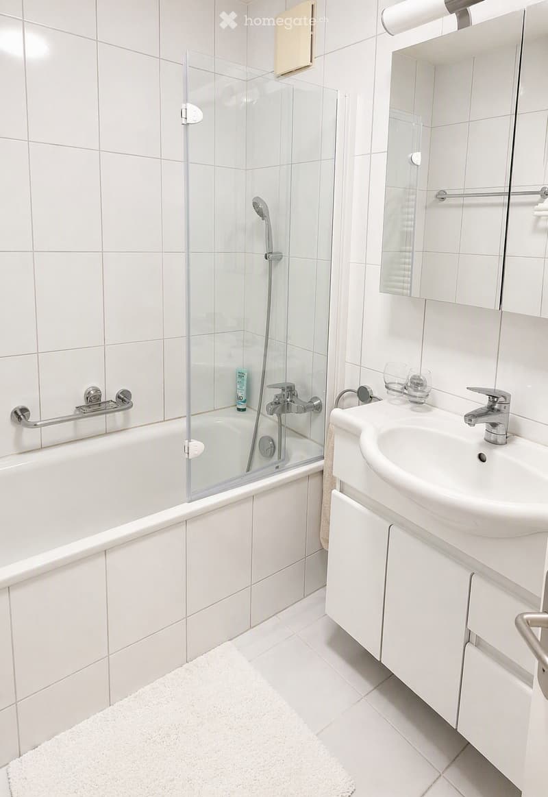 Modern bathroom with white tiles, a bathtub with a glass shower enclosure, a sink with a mirror, and a small white rug on the floor.