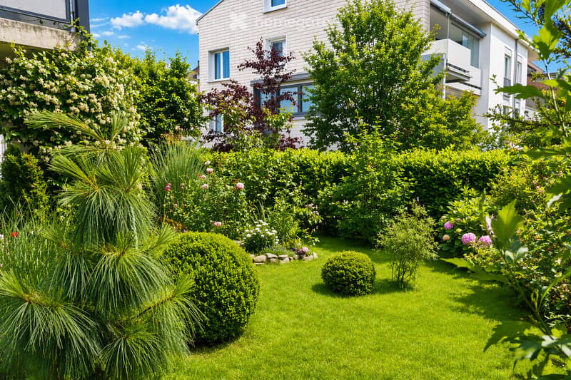 Well-maintained garden with green lawn, trimmed bushes, flowering plants, and trees, with a modern house in the background.