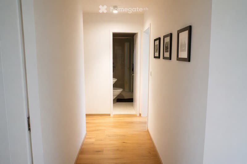 A clean, well-lit hallway with light wooden flooring and white walls. Three framed pictures are mounted on the right wall, and an open doorway leads to a bathroom with a toilet, sink, and bathtub.