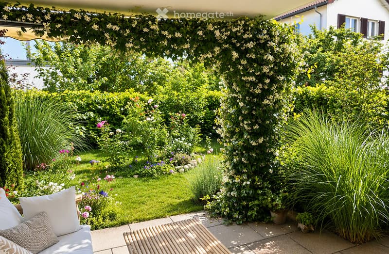 A well-maintained garden with a variety of flowering plants, shrubs, and tall grasses. A white outdoor sofa with cushions and a wooden bench are placed on a paved patio area.