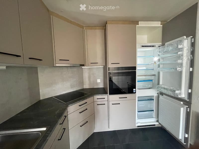 Modern kitchen with light-colored cabinets, black countertops, and stainless steel appliances including a refrigerator and oven. The refrigerator door is open, revealing its interior.
