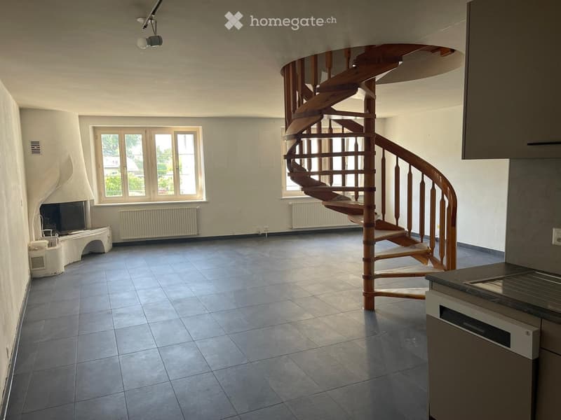 Open-plan space with a spiral wooden staircase, tiled floor, large windows, kitchen counter, radiator, and fireplace.