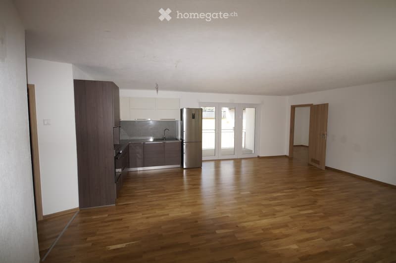 Empty room with wooden flooring, white walls, and a kitchen area featuring cabinets, a sink, and a refrigerator. The room has large glass doors leading to an outdoor area.
