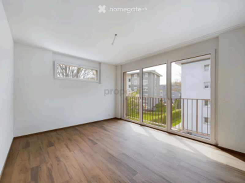 Bright, unfurnished room with wooden flooring, large sliding glass doors leading to a balcony, and a small window on one wall.
