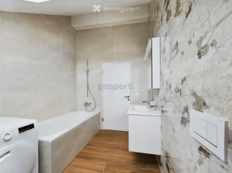 Modern bathroom with a white bathtub, sink, mirror, and washing machine. The walls are a mix of light grey and exposed stone, and the floor is wooden.