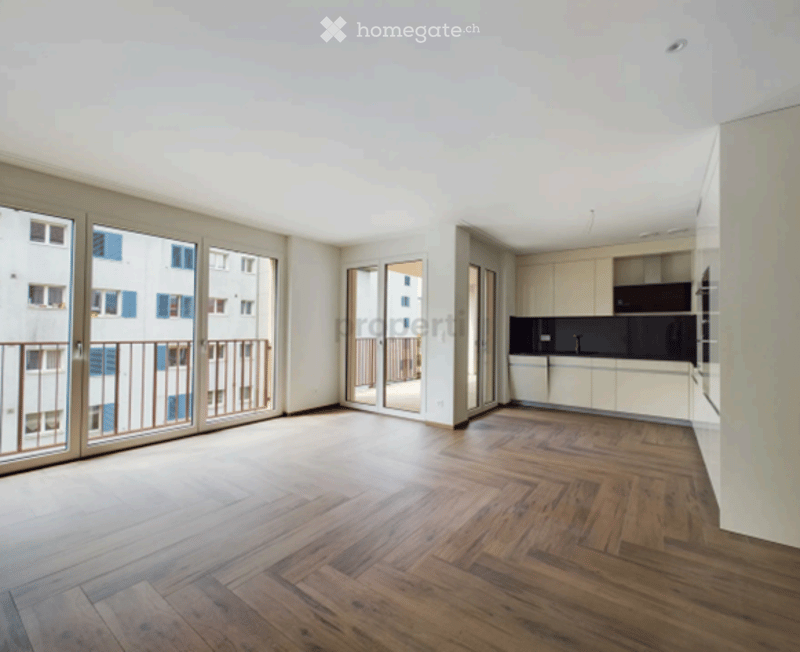 Bright, unfurnished living space with large glass doors leading to a balcony, wooden herringbone flooring, and a modern kitchen area with cabinets and appliances visible in the background.