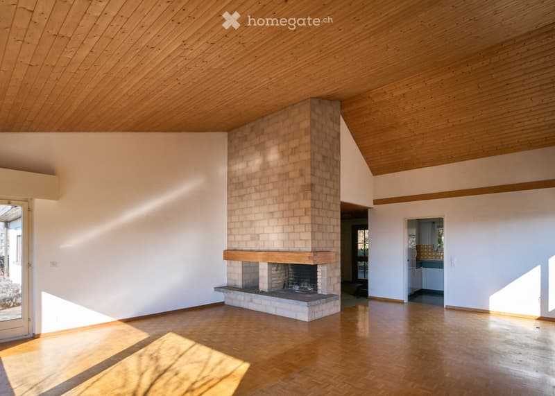 Large open room with wooden flooring, tall wooden ceiling, central brick fireplace with mantle, sliding door to the garden, white walls