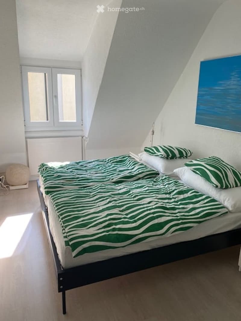 Spacious bedroom with a double bed, white and green bedding, white pillows, double windows, white walls, wooden floor, a picture on the wall, and a white radiator