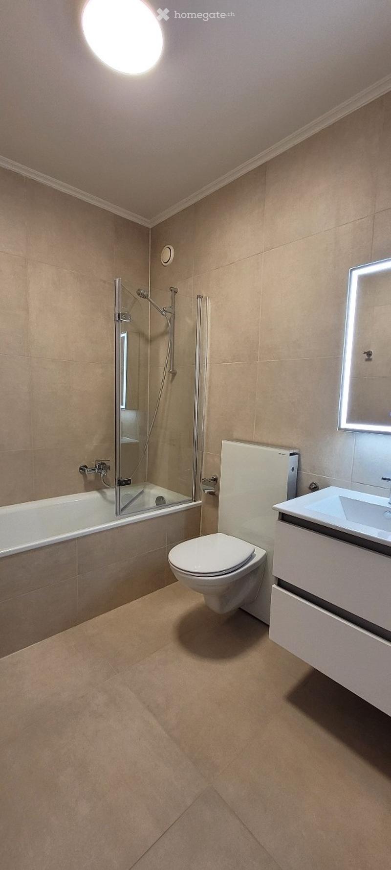 Modern bathroom with beige tiles, a bathtub, toilet, sink, and a glass-enclosed shower area.