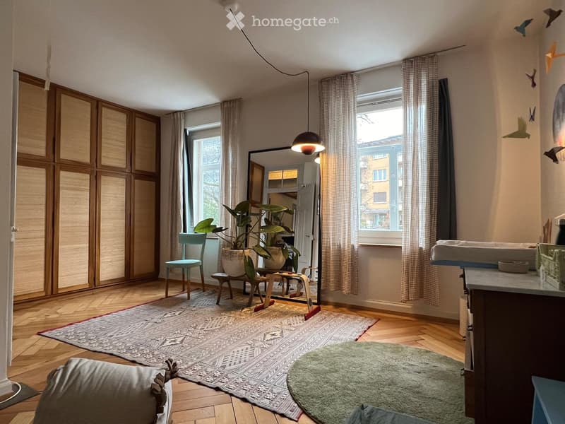 A modern, well-lit room with wooden flooring, large windows, and a mix of contemporary and minimalist furniture including a sofa, chair, table, and potted plants.