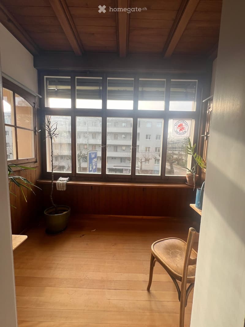 Empty room with wooden flooring, large windows, and potted plants on the windowsill. A single wooden chair is placed near the window.