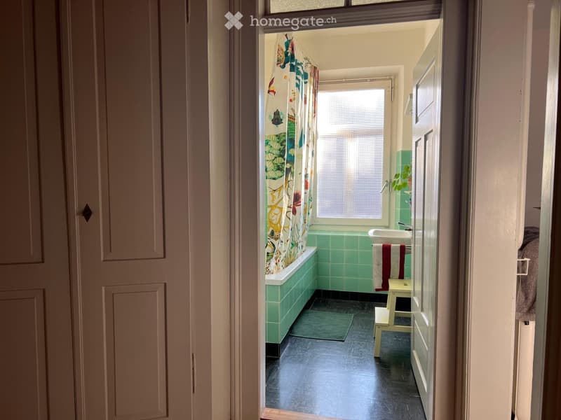 Bathroom with green tiled walls, a bathtub with a colorful shower curtain, a sink, a small stool, and a towel hanging on the door.