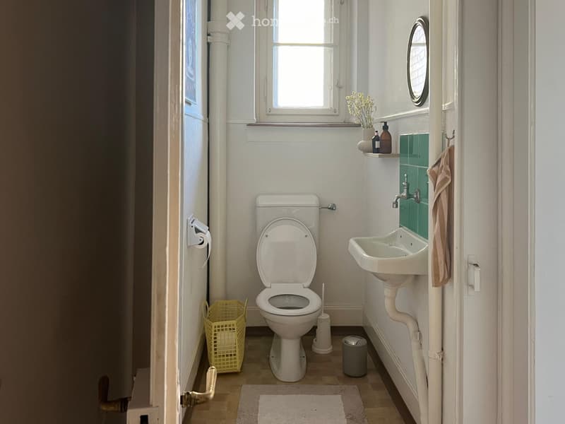 A small, well-lit bathroom with a toilet, sink, mirror, and a small window. Includes a towel, toilet paper holder, flower vase, and a basket on the floor.