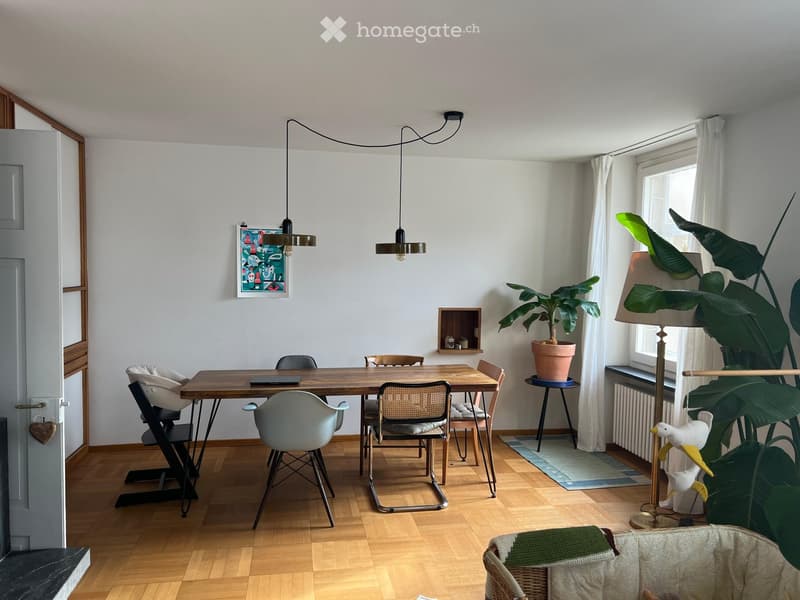 Bright, modern room with wooden flooring, a large wooden dining table surrounded by chairs, a baby chair, a potted plant, a floor lamp, and a decorative picture frame on the wall.