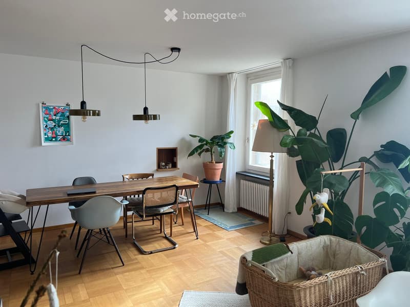 Bright living space with wooden flooring, large window with white curtains, modern pendant lights, dining table with chairs, floor lamp, potted plants, and a wicker basket with toys.