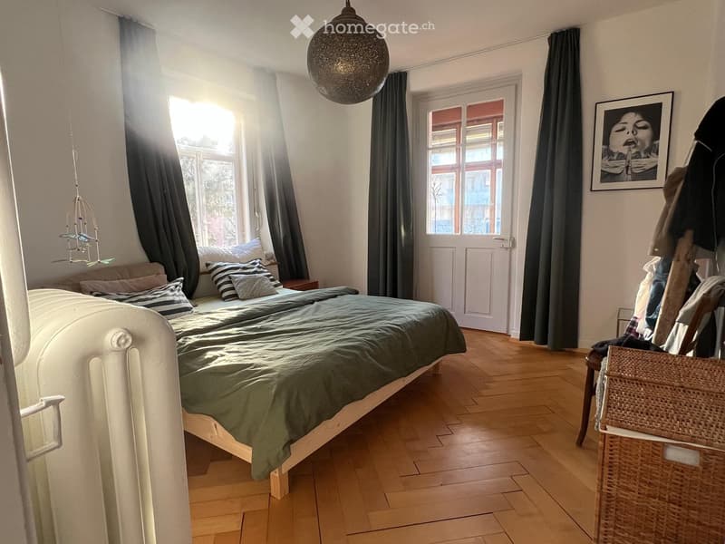 Cozy bedroom with wooden flooring, a white bed with green bedding, nightstand, picture frame, and a hanging lamp.