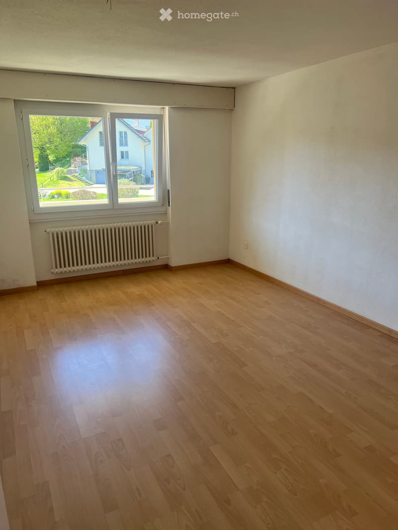 Empty room with wooden flooring, white walls, a radiator, and a large window offering a view of the outside garden and neighboring house.