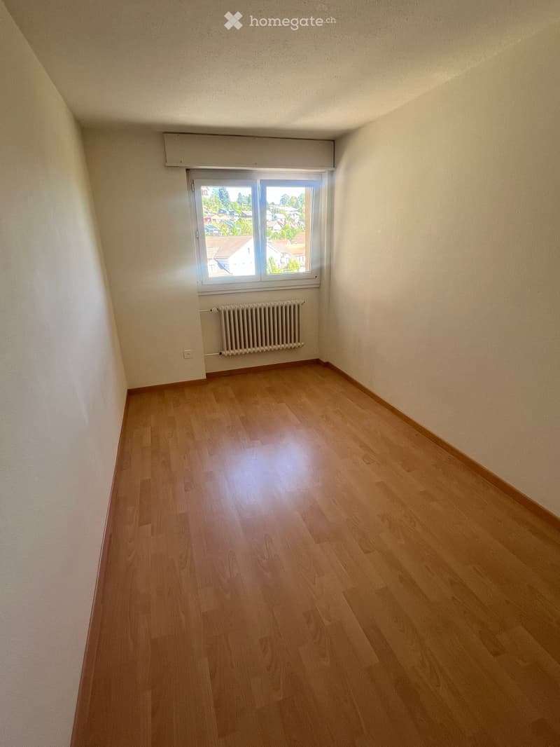 Empty room with wooden flooring, white walls, a window with a view of rooftops, and a radiator below the window.