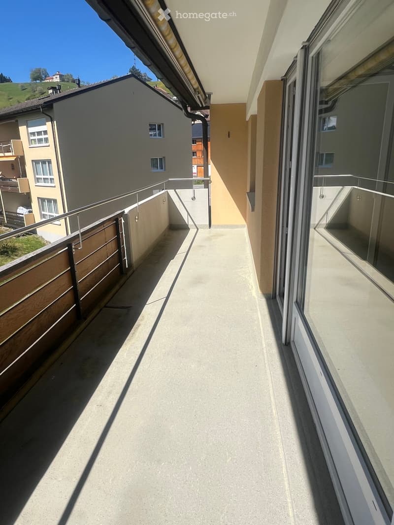 Balcony with concrete floor, metal railing, and wooden paneling, overlooking a residential area with houses and greenery.