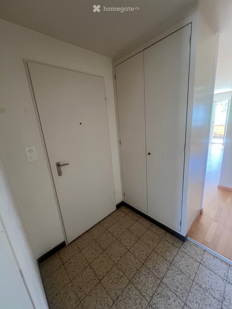 Empty hallway with tiled floor, white walls, and a closed door on the left. Natural light comes from an open doorway on the right leading to another room with wooden flooring.