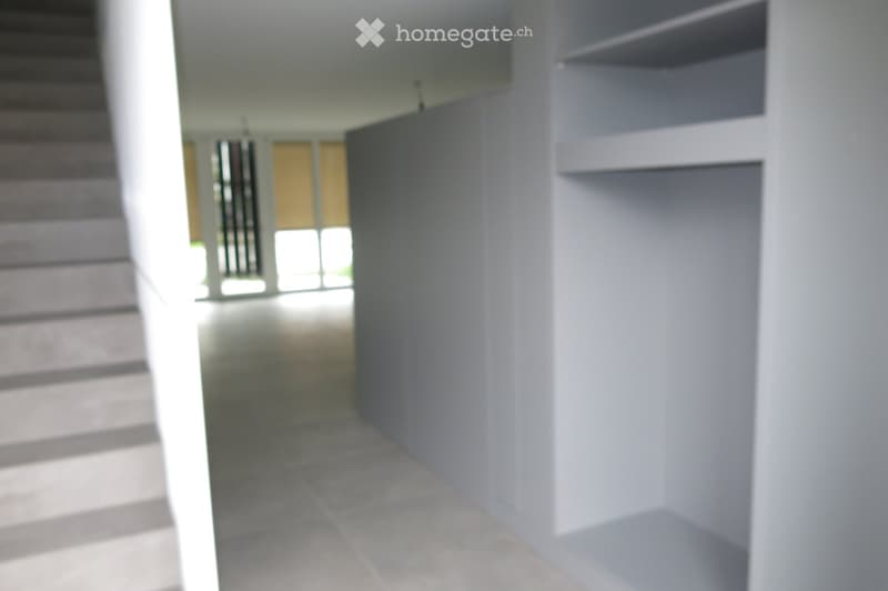 Empty hallway with white walls, tiled floor, and a staircase leading upwards. The area is well-lit with natural light coming from a window at the end of the corridor.