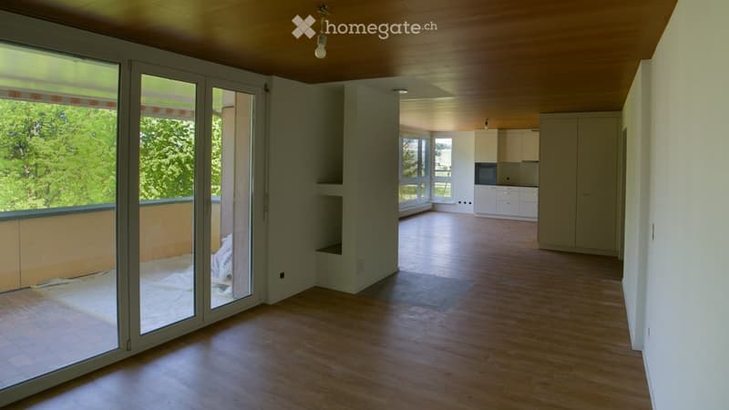 Empty living space with wooden flooring, large sliding glass doors leading to a balcony, and a view of greenery. The adjacent kitchen area features white cabinets and a countertop.