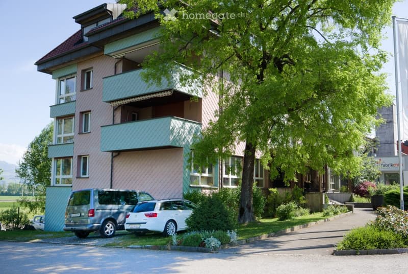 Two-story building with balconies, multiple windows, and a parking area with two cars.