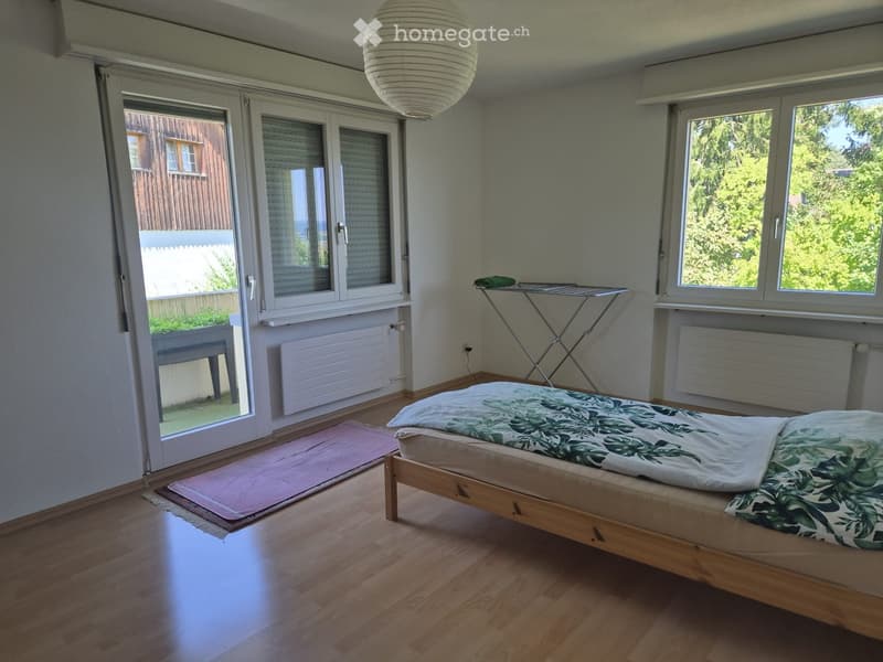 Bright bedroom with wooden flooring, large windows, a bed with green patterned bedding, a radiator, a chair, and an ironing board.