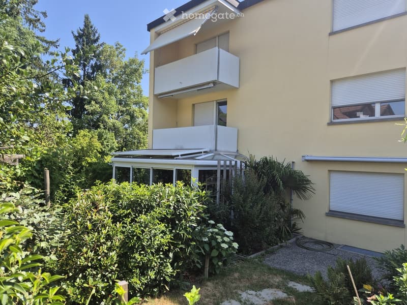 Two-story building with light yellow walls, multiple windows with roller blinds, and balconies with white railings.