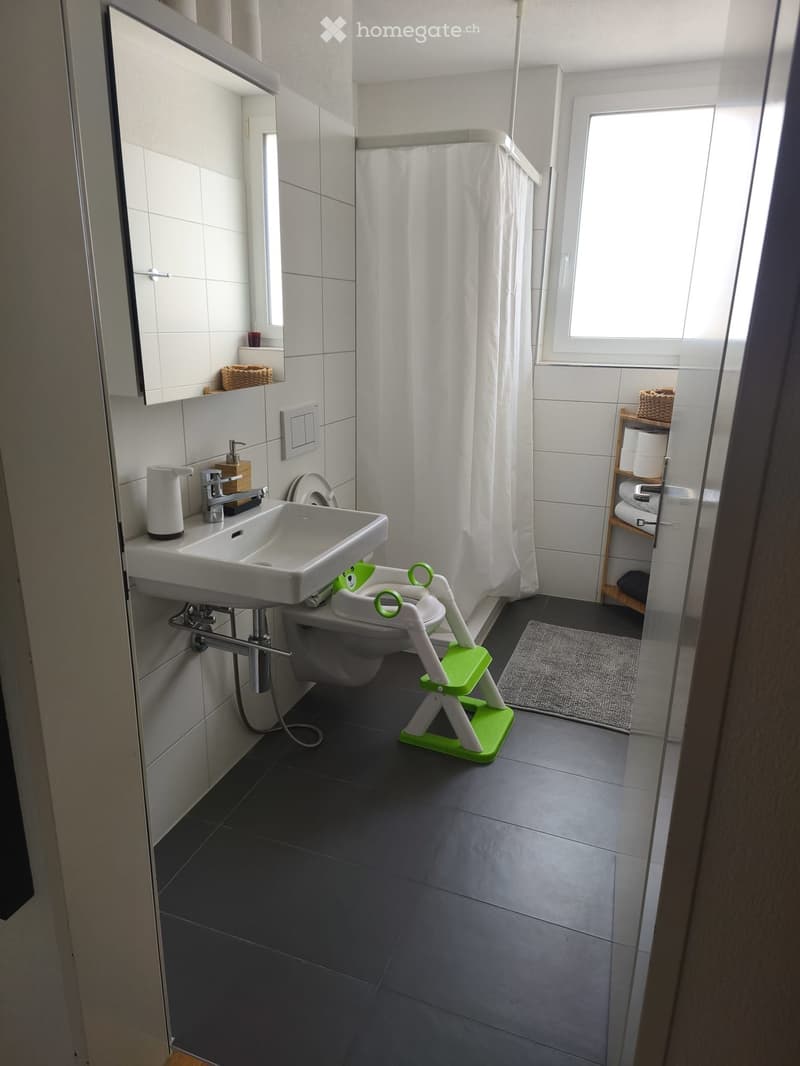 Modern bathroom with white tiles, a sink, toilet, shower curtain, and a small green step stool for children.