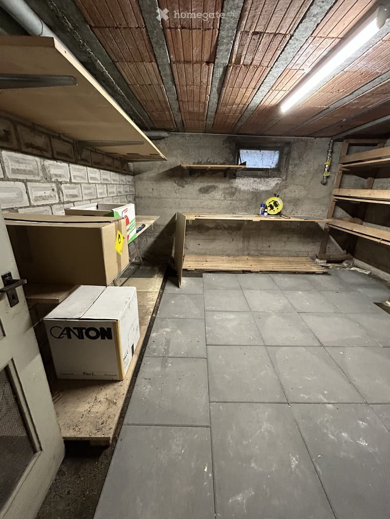 Empty basement or utility room with concrete walls, tiled floor, exposed wooden beams, and some storage boxes and shelves.