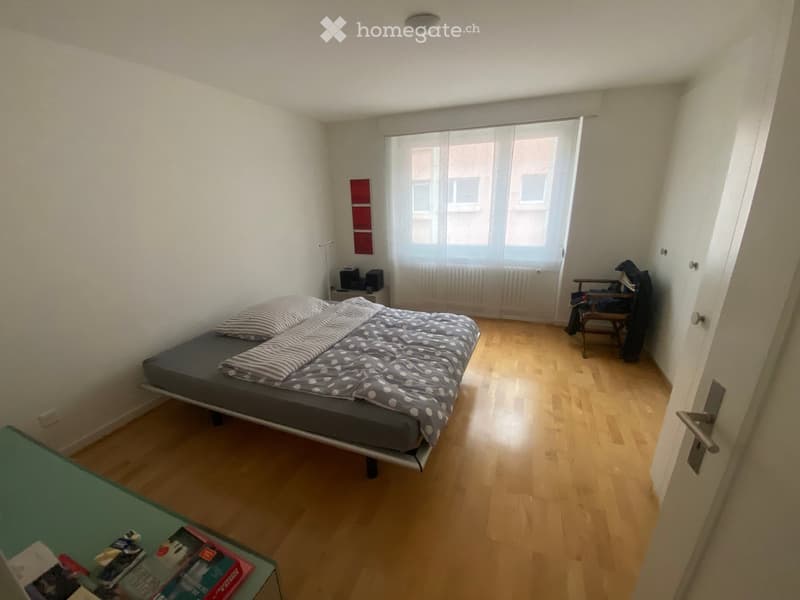 A small, furnished bedroom with a sofa bed, a wooden chair, a small table with books, a lamp, and a radiator near the window.