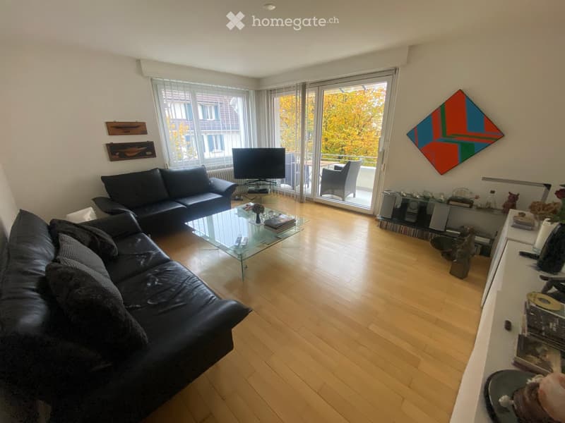 Modern living room with wooden flooring, large windows, and a balcony. Features a black leather sofa, glass coffee table, television, and decorative art pieces.