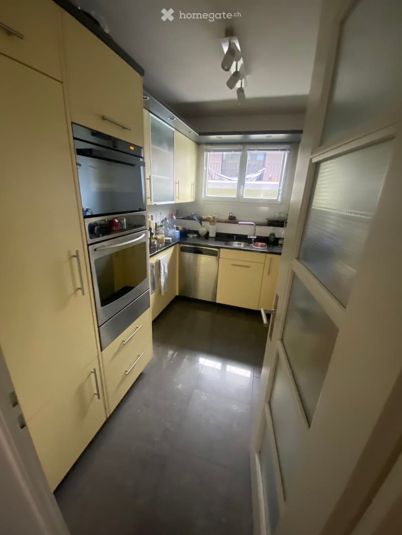 Modern kitchen with light-colored cabinets, stainless steel oven, dishwasher, and sink, featuring dark tiled flooring and ample natural light from windows.