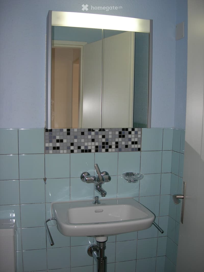 Bathroom with a white sink, chrome faucet, and a mirrored cabinet with a built-in light above it. The walls are tiled in light blue with a decorative mosaic border near the mirror.