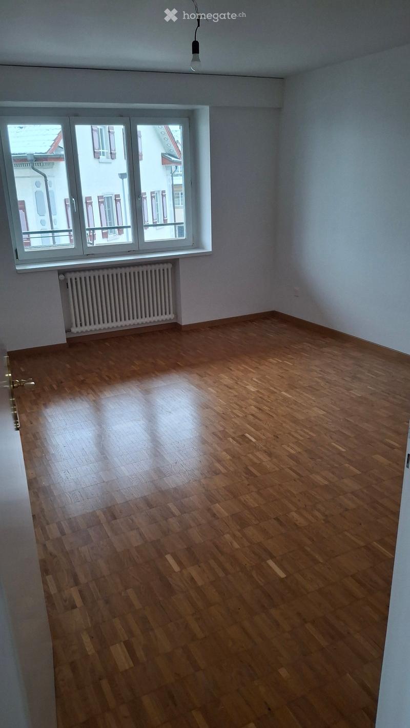 Empty room with wooden flooring, white walls, and a large window with a radiator below it.