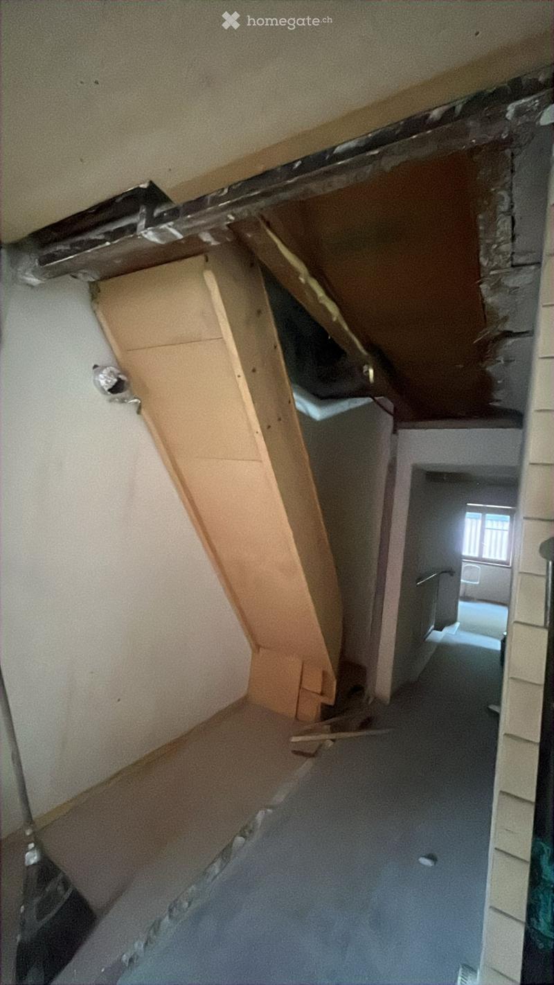A narrow, unfinished hallway with exposed wooden beams, a partially removed ceiling, and a visible doorway leading to another room.
